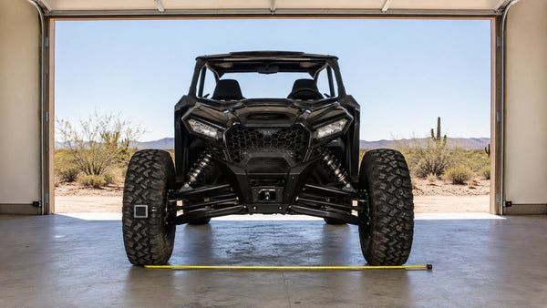 Sport side-by-side UTV parked head-on in an open garage on level concrete, with a tape measure laid across the ground between the front tires to check toe alignment
